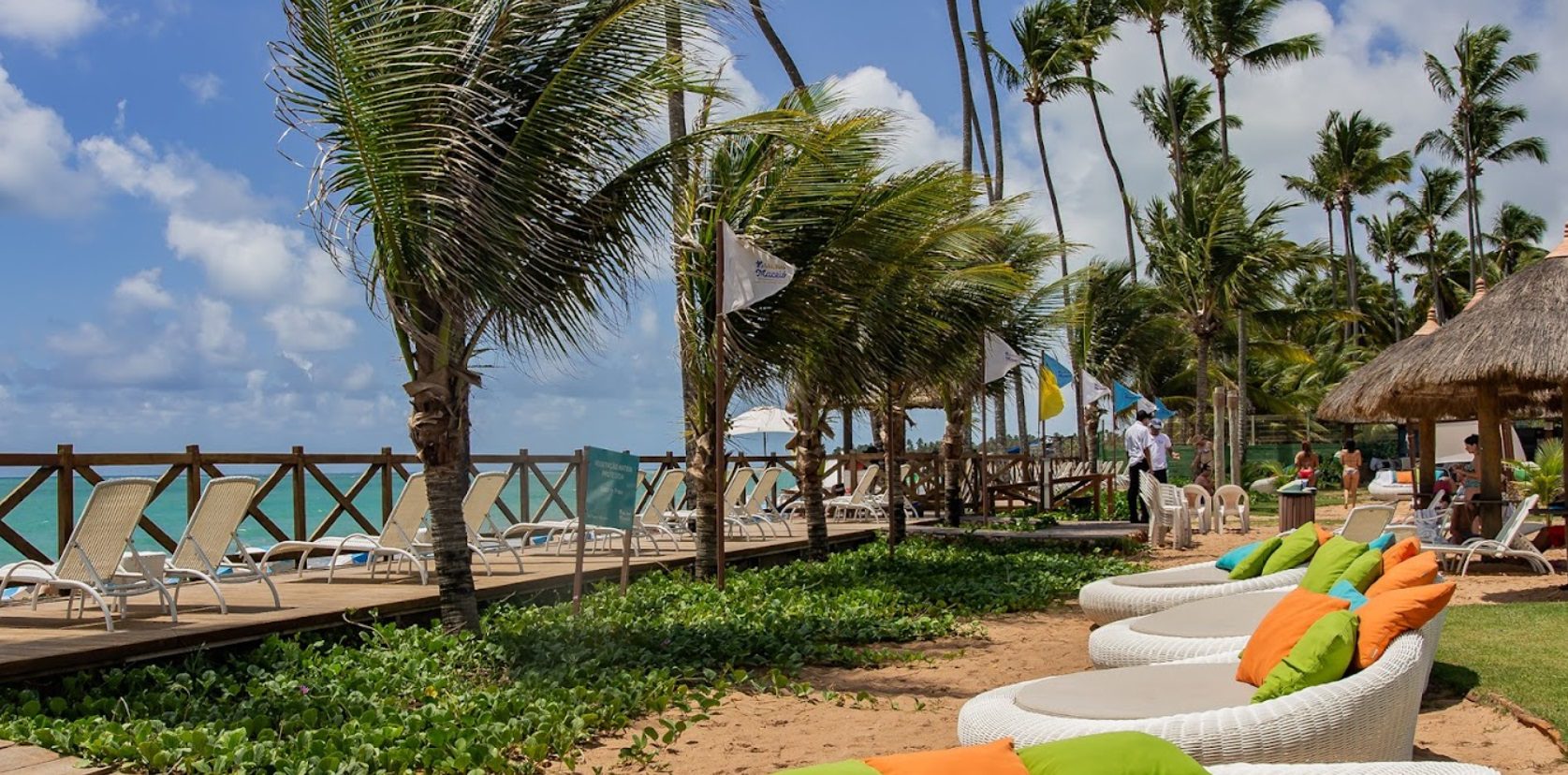Vista-de-um-resort-à-beira-mar-Salinas-Maceió-All-Inclusive-Resort-Maceió-AL Vista de um resort à beira-mar com espreguiçadeiras brancas ao longo de uma passarela de madeira, cercada por palmeiras. O mar cristalino e o céu claro criam um ambiente relaxante e tropical.