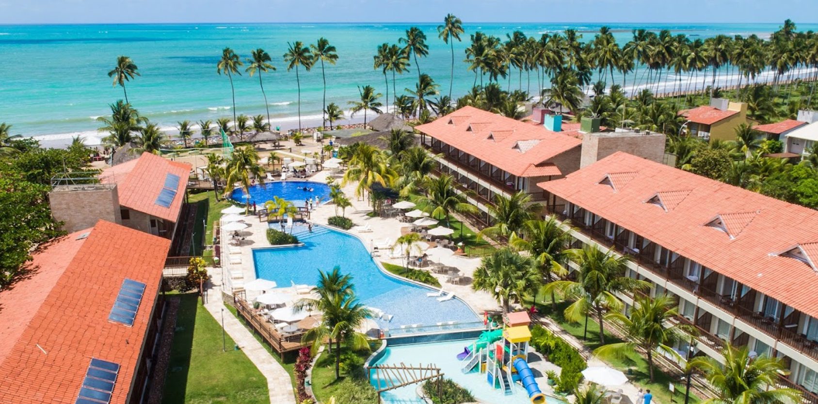resort-beira-mar-com-palmeiras-ao-redor-Salinas-Maceió-All-Inclusive-Resort-Maceió-AL Imagem aérea de um resort à beira-mar, com palmeiras ao redor e uma grande piscina central. Ao fundo, a praia com águas cristalinas, enquanto no primeiro plano, um toboágua colorido e espreguiçadeiras oferecem conforto aos hóspedes.