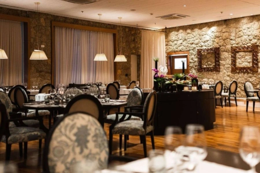 restaurante elegante dentro de um hotel resort. O ambiente é sofisticado, com paredes de pedra exposta e iluminação suave proporcionada por pendentes de tecido. As mesas estão dispostas com talheres e copos, e as cadeiras possuem estofados florais. No fundo, há grandes janelas com cortinas brancas e alguns detalhes decorativos, como espelhos e plantas, que complementam o ambiente acolhedor e requintado.