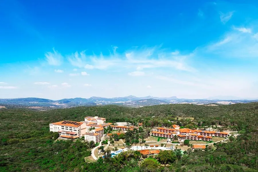 Vista panorâmica de um resort cercado por vastas áreas verdes e montanhas ao fundo, sob um céu azul claro. O complexo de edifícios com telhados vermelhos se destaca na paisagem natural.