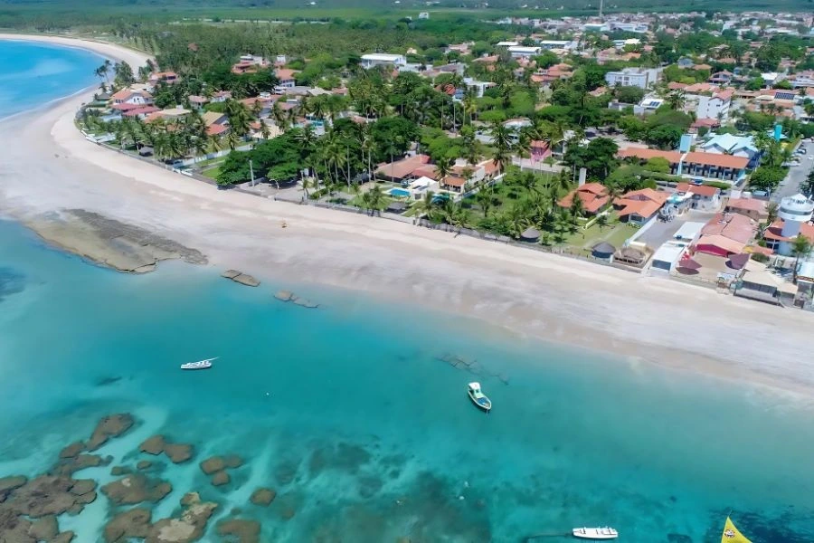 aérea de uma praia com águas cristalinas e rasas, destacando o trecho em frente ao Summerville All Inclusive Resort. A praia é cercada por casas e estabelecimentos à beira-mar, com palmeiras e vegetação ao longo da costa. Há também algumas rochas visíveis na água, criando uma paisagem tranquila e natural. Um barco pequeno está flutuando nas águas calmas. O resort, com seus telhados vermelhos, é visível à direita da imagem, com uma piscina na parte inferior.