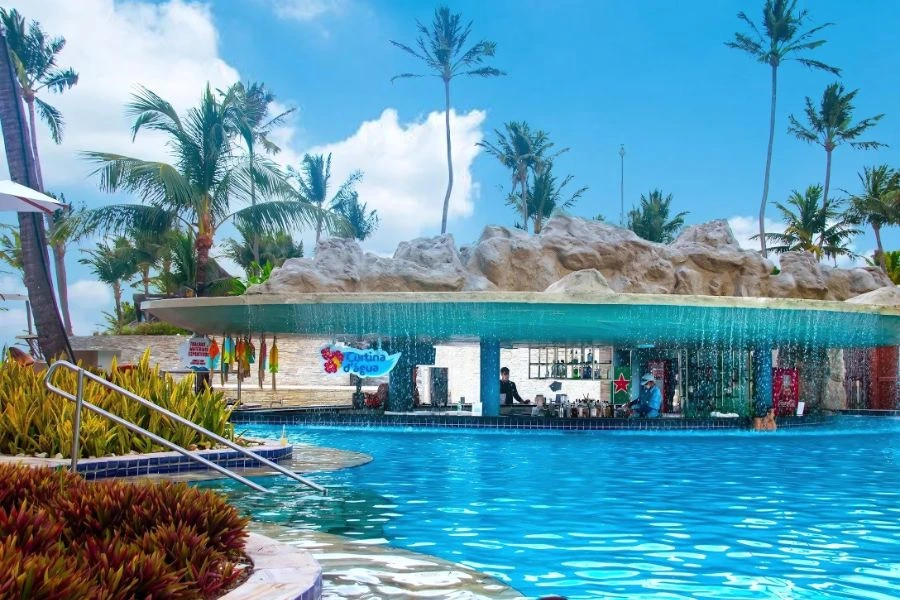 Imagem de uma piscina com um bar submerso, onde uma cortina de água cai sobre o local. O bar é coberto por uma estrutura de pedras, criando um ambiente natural e relaxante. Ao fundo, é possível ver um bartender trabalhando, enquanto os hóspedes desfrutam da piscina. Palmes iluminam o espaço ao redor, com vegetação tropical que completa a cena, enquanto o céu está claro e ensolarado, criando uma atmosfera de férias paradisíacas.
