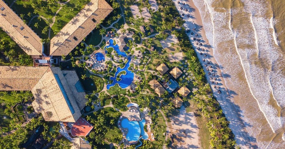 area-externa-superior-terreno-a-beira-mar-Transamerica-Comandatuba-Una-BA Imagem aérea de um resort à beira-mar, com a praia se estendendo ao longo da costa. O complexo é rodeado por uma grande área verde e várias palmeiras, criando um ambiente tropical. O resort conta com diversas piscinas, espreguiçadeiras e áreas de lazer. À direita, um rio se encontra com a praia, e é possível ver algumas embarcações flutuando na água. O céu está claro e o sol ilumina a paisagem, proporcionando uma atmosfera relaxante e paradisíaca. Ao fundo, há mais vegetação e áreas residenciais.