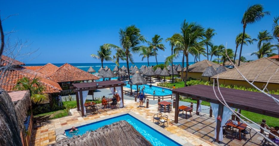 imagem-resort-Ocaporã-Hotel-All-Inclusive-piscina-guarda-sol-mesas-cadeiras-a-beira-mar Imagem de um resort à beira-mar, com várias cabanas de palha ao redor de piscinas de formato orgânico. As espreguiçadeiras estão dispostas ao redor das piscinas, e há pequenas áreas com mesas e cadeiras de madeira sob pérgulas. Ao fundo, o mar azul e calmo se estende, com o céu limpo e ensolarado. O ambiente é rodeado por palmeiras, criando uma atmosfera tropical e relaxante, perfeita para um dia de lazer à beira da praia.