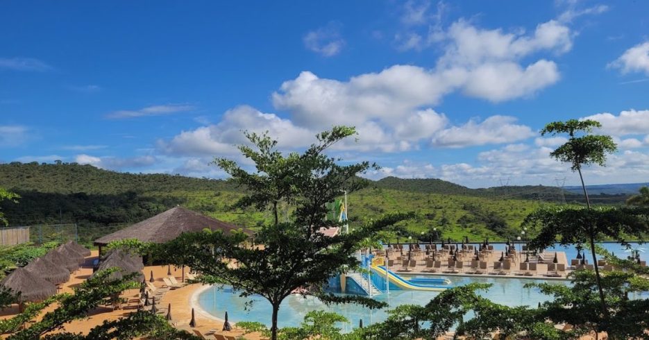resort-taua-alexania-GO-área de lazer-piscina-toboágua-colorido Imagem de um resort com uma grande área de lazer, destacando uma piscina com toboágua colorido. Ao redor da piscina, há várias espreguiçadeiras e sombrinhas, além de palmeiras e vegetação tropical que embelezam o espaço. O cenário é rodeado por colinas verdes e um céu claro, com poucas nuvens, criando uma atmosfera tranquila e relaxante. A arquitetura do resort é simples e funcional, oferecendo um ambiente de lazer e bem-estar.