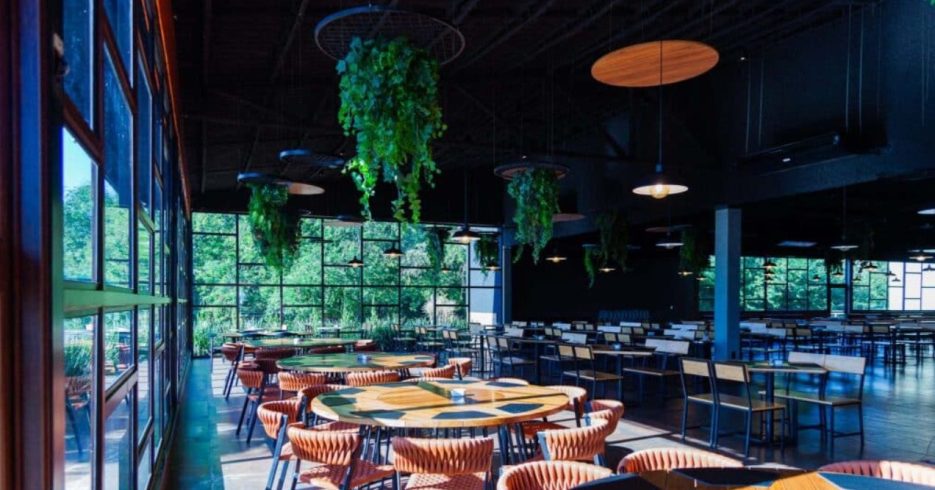 restaurante-Tauá-Convention-Atibaia-SP interior de um restaurante moderno e arejado, com mesas circulares e cadeiras de design contemporâneo. O ambiente possui grandes janelas que oferecem uma vista para a vegetação externa, iluminando o espaço com luz natural. Pendentes de luz e plantas suspensas no teto adicionam um toque de frescor ao ambiente. O estilo minimalista e a combinação de cores neutras criam uma atmosfera acolhedora e relaxante para os clientes.