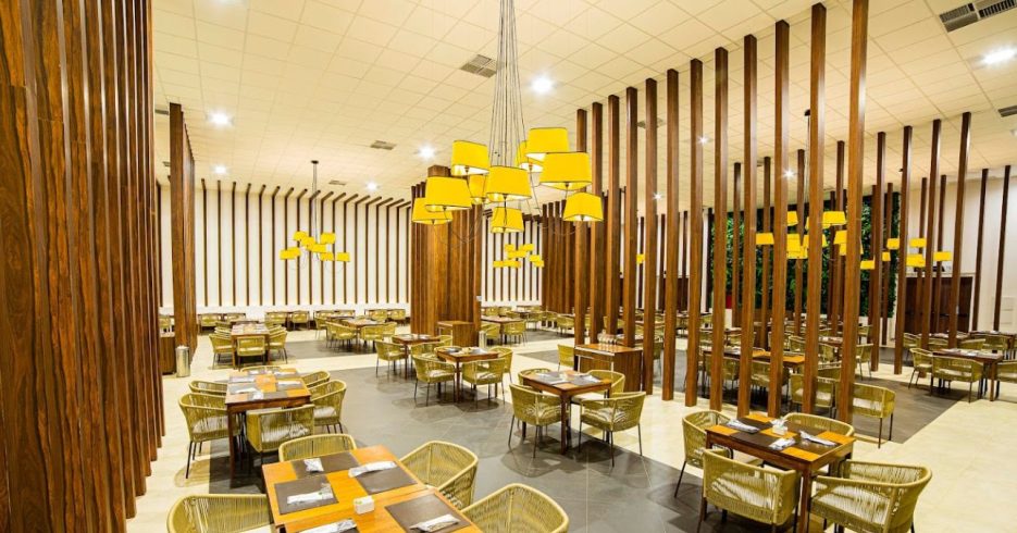 restaurante-resort-Tauá-Resort-Convention-Alexânia-GO Imagem do interior de um restaurante moderno, com mesas dispostas de maneira organizada. O espaço é decorado com colunas de madeira vertical que criam uma sensação de profundidade, além de luminárias amarelas que pendem do teto, proporcionando uma iluminação suave. As cadeiras de vime e os arranjos nas mesas adicionam um toque aconchegante ao ambiente. No fundo, há uma parede com vegetação, contrastando com o design elegante e minimalista do restaurante. O ambiente é amplo e bem iluminado, criando uma atmosfera acolhedora e moderna.