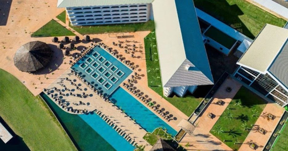 visão-superior-Tauá-Resort-Convention-Alexânia-GO Imagem aérea do Tauá Resort & Convention – Alexânia (GO) com uma grande área de piscina, cercada por espreguiçadeiras e guarda-sóis. A piscina tem um design moderno, com formas retangulares interligadas. Ao fundo, duas grandes construções do resort são visíveis, com telhados de metal e paredes claras. O ambiente é bem organizado, com jardins e áreas verdes ao redor, criando um espaço relaxante e convidativo. A iluminação natural e o céu limpo completam a cena, proporcionando uma atmosfera tranquila.