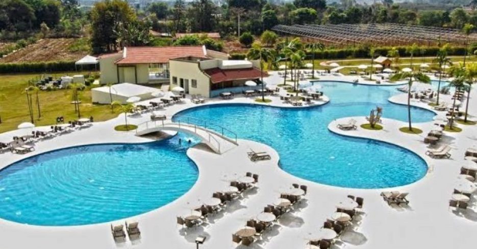 vista-aerea-superior-do-piscina-Tauá-Resort-Convention-Atibaia-SP grande complexo de piscinas com formas orgânicas e interligadas. Ao redor das piscinas, há várias espreguiçadeiras e guarda-sóis, proporcionando uma área de relaxamento para os hóspedes. O local é rodeado por vegetação e ao fundo é possível ver uma construção de telhado vermelho, possivelmente um restaurante ou centro de apoio. O ambiente é bem iluminado, com céu claro e ensolarado, criando uma atmosfera de lazer e tranquilidade.