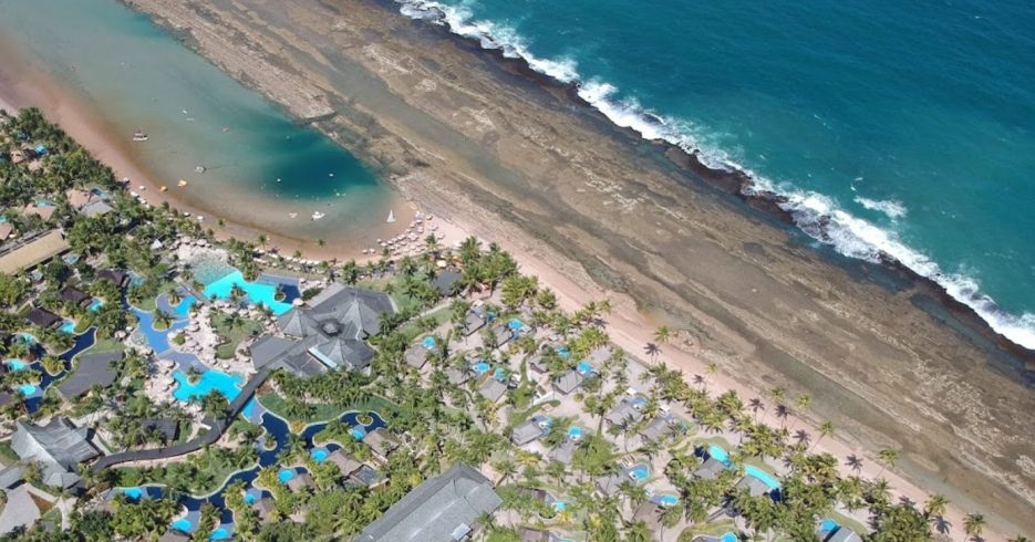 vista-aerea-superior-do-terreno-Nannai-Resort-Spa-Porto-de-Galinhas-PE Imagem aérea de um resort à beira-mar, com várias piscinas interligadas e construções espalhadas pela área, todas rodeadas por vegetação tropical. A piscina principal apresenta formas orgânicas e algumas áreas de descanso com espreguiçadeiras. À direita, o mar azul e claro é visível, com uma faixa de areia ao longo da costa e algumas embarcações ao longe. A imagem transmite uma sensação de tranquilidade e lazer, destacando o ambiente natural e a estrutura do resort.