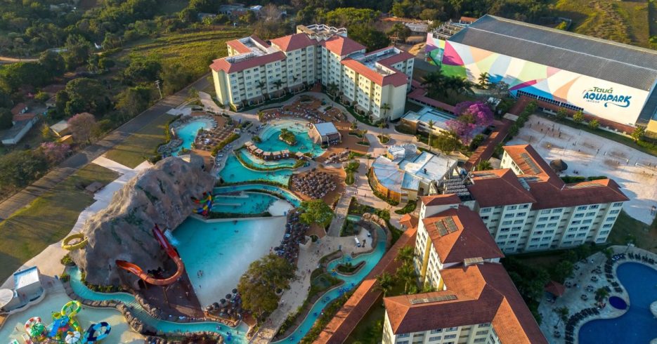 vista-aerea-superior-do-terreno-Tauá-Resort-Convention-Atibaia-SP Imagem aérea de um resort com um grande parque aquático ao centro. O complexo conta com várias piscinas, toboáguas coloridos, e uma grande estrutura de rochas artificiais com cachoeiras. As espreguiçadeiras estão dispostas ao redor das piscinas e a área verde do resort se mistura com o ambiente de lazer. Ao fundo, é possível ver o edifício principal do resort e um grande pavilhão com a inscrição "Tauá Aquapark Indoor". A paisagem é rodeada por árvores e vegetação, proporcionando um ambiente natural e relaxante.